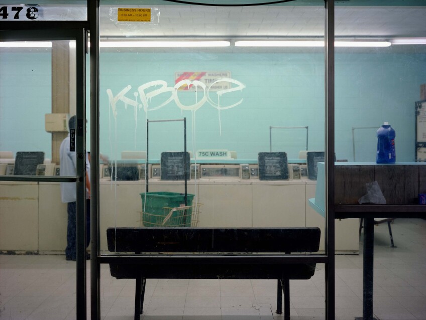 A laundromat taken from the outside. The floor to ceiling windows provide a complete view of the laundromat with a light blue wall. A bench is pushed up against one of the windows. The window has graffiti on it, reading "KBOE." A man is seen standing in front of one of the washing machines.
