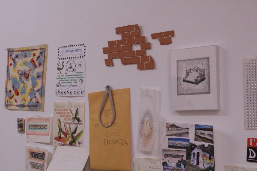 A wall is covered with various art pieces from prints and photographs to flat sculpture pieces like a mosaic of flattened brick lined with white. 