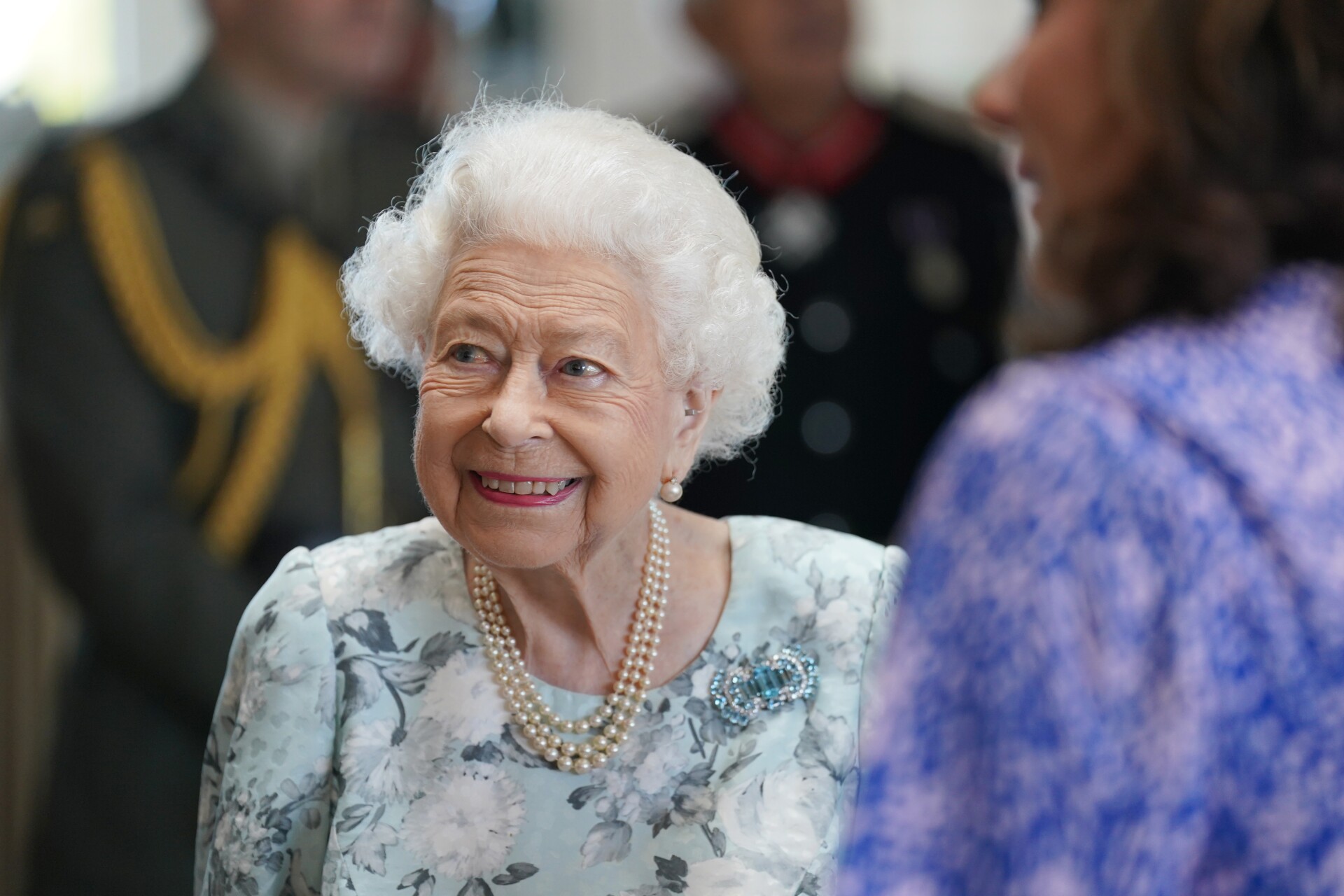 Queen Elizabeth II smiles while looking towards her right, wearing a blue floral dress
