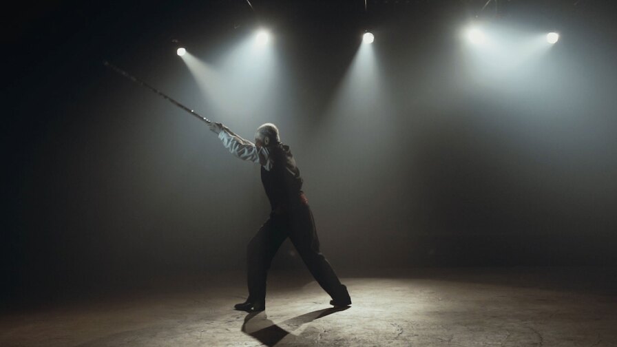 Still from Border Blaster Video: Person on a stage with spotlights thrusts a long object into the air