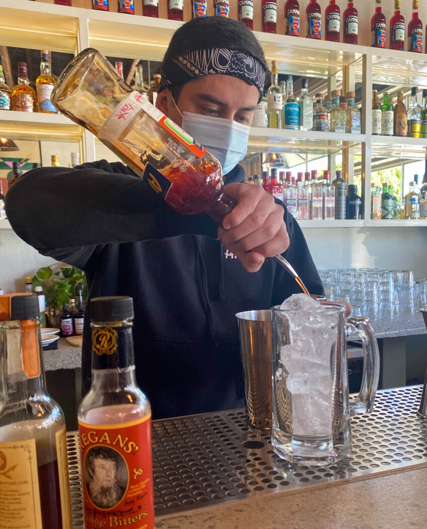Kevin Gomez, a man with a bandana tied around his forehead and a PPE face mask over his nose and mouth stands behind a bar counter and pours a red liquor into a tall glass filled with ice. Behind him are shelves full of liquor bottles.