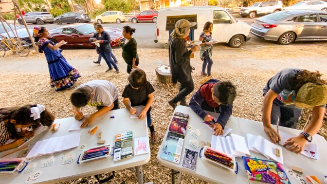 Participants make zines on outdoor tables. Behind them, people are standing around and eating food.