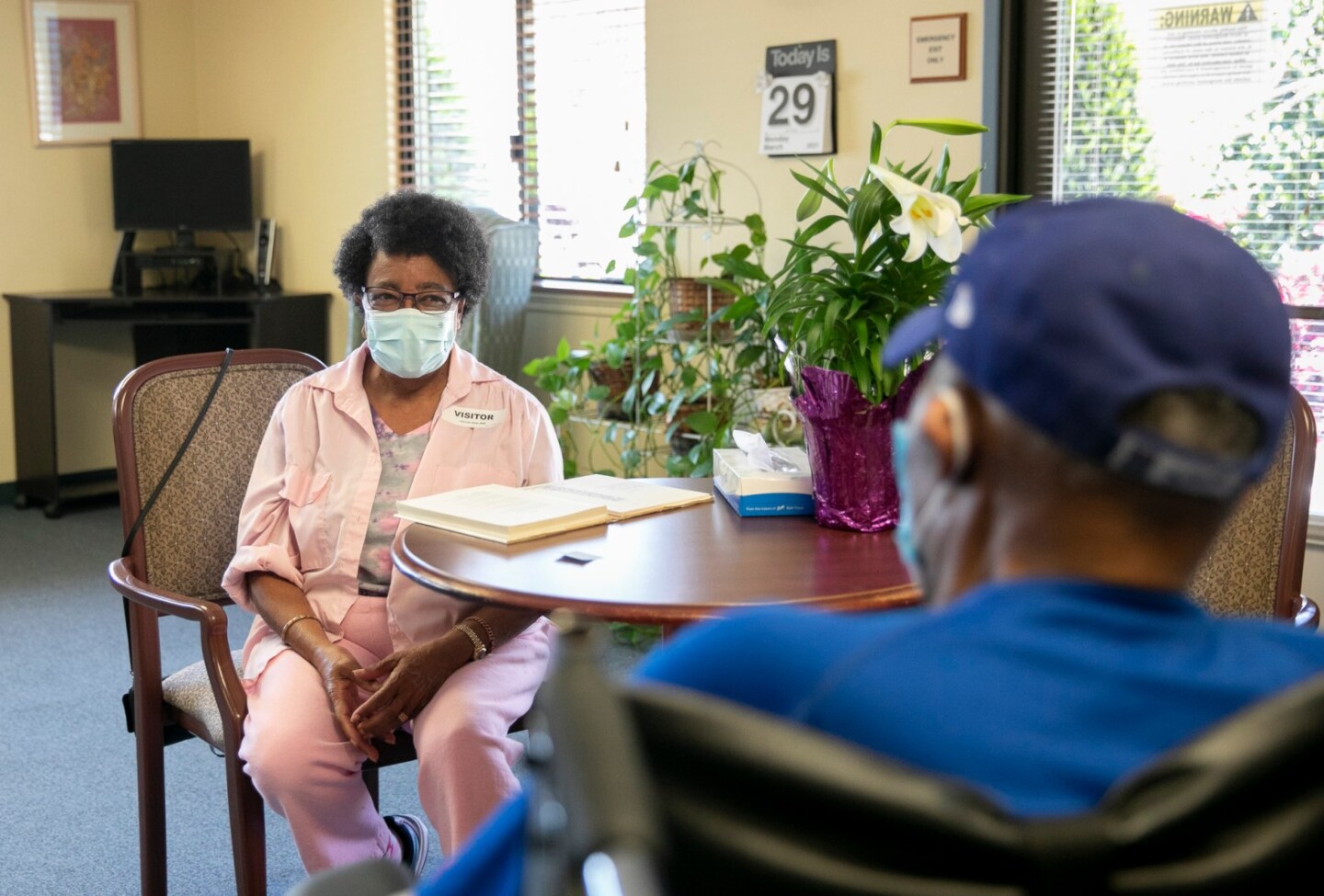 A Black woman wearing a face mask and a pink jacket and pants looks longingly at a man in a baseball cap sitting across her inside a nursing home.