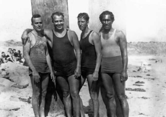 A black and white photo of (from left to right) Gerard Vultee, Owen Hale, Bill Herwig and Duke Kahanamoku at a beach. The men are standing in front of two wooden surfboards propped up on the sand.