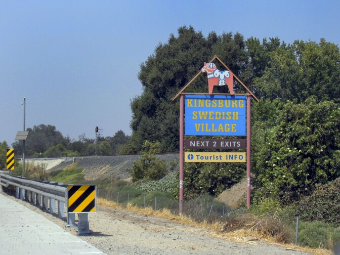 A sign along the side of a road reads, "Kingsburg Swedish Village" in yellow lettering over an electric blue background. Beneath that reads, "Next 2 exits" and below that reads, "Tourist INFO." Above the signage is an icon of a red horse with a white saddle.