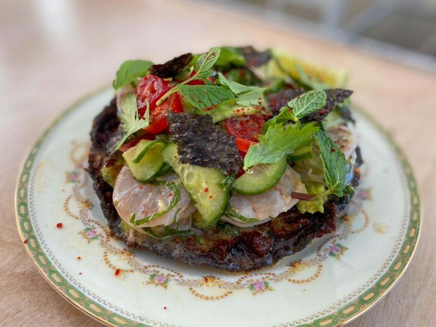 Shrimp ceviche sits over a black tostada and is topped with chopped cucumber, red peppers, fresh green herbs and seaweed.