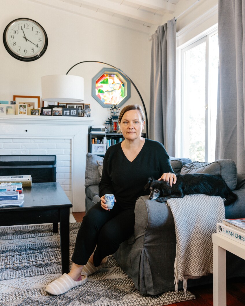 Mila Inukai sits with her dog in her living room. The room is dressed with a large clock, photos and a bookshelf.