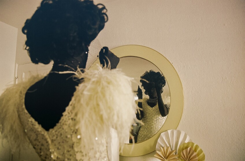 Muxxxe, who is wearing a black morph suit underneath a white sparkly dress, looks in the mirror and adjusts their wig which sits on top of their head.