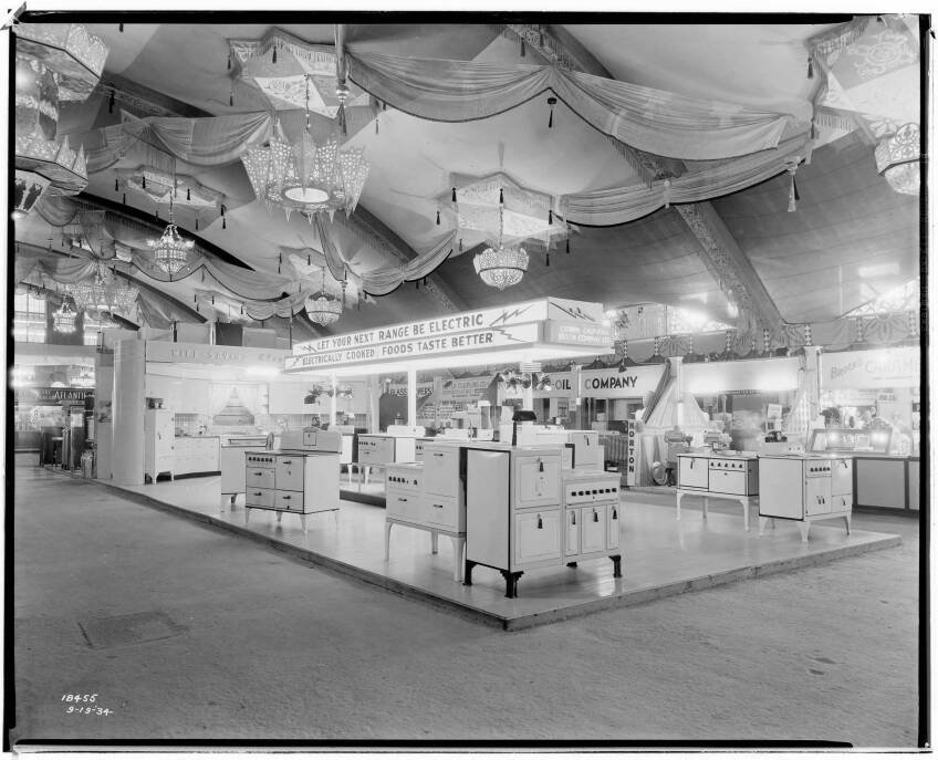 A black and white photo of an electricity exhibit at the Los Angeles County Fair. Various appliances are arranged on a showroom floor and lit with bright lights. A sign that reads, "Let your next range by electric. Electrically cooked foods taste better" stands above the cooking ranges below.