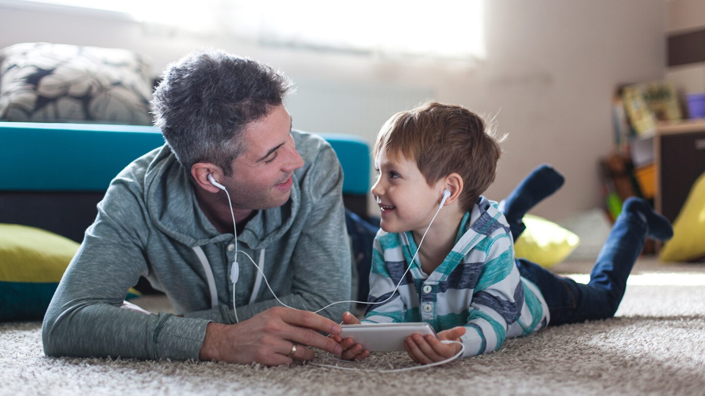 A man and a little boy watch something on a tablet together as they lie down on a carpet next to each other