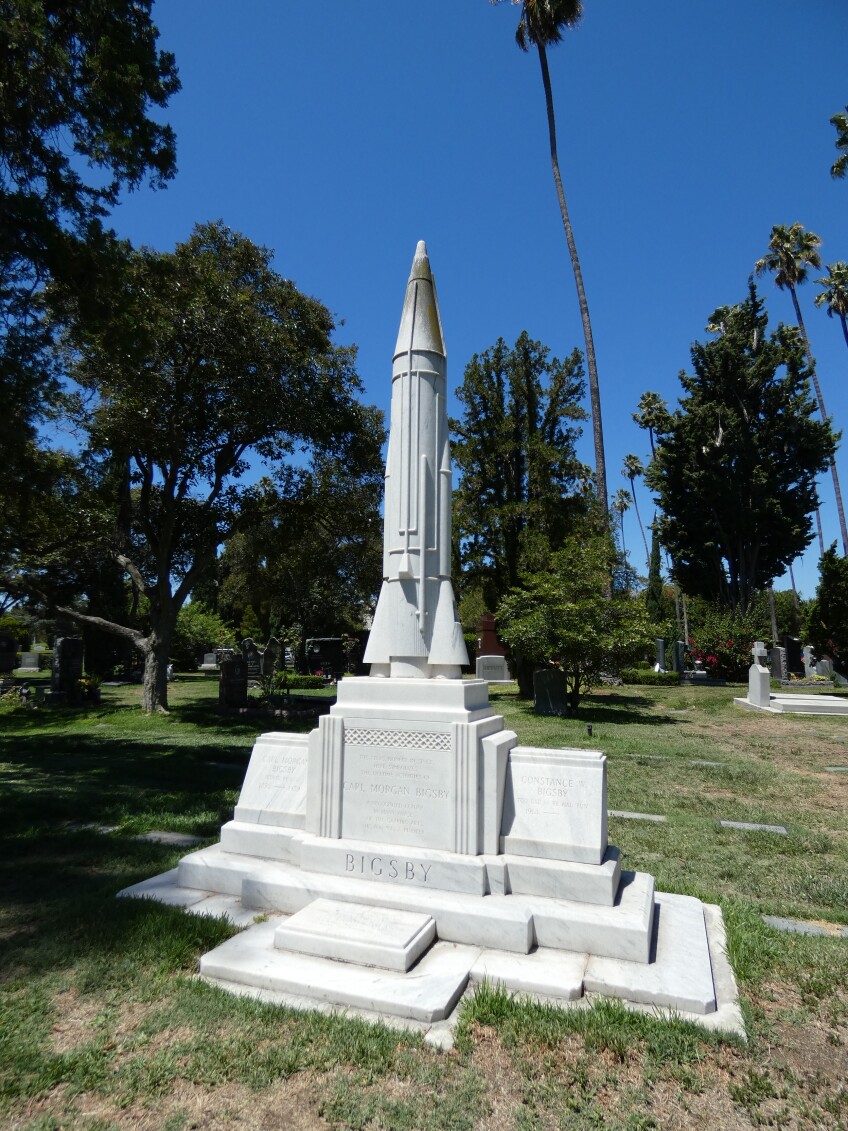 A white marble tomb shaped like a rocket stands on a white marble pedestal. At the base of the pedestal is engraved, "Bigsby." 