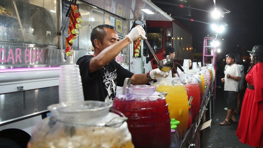 Fruit juices in jugs are lined up on a table in front of a food truck. A man in a black t-shirt uses a ladle to scoop juice into a small sample cup for a customer.