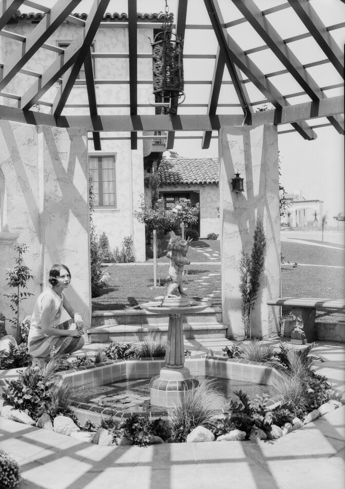 A woman poses beneath a gazebo with a fountain and a beautiful house in the background.