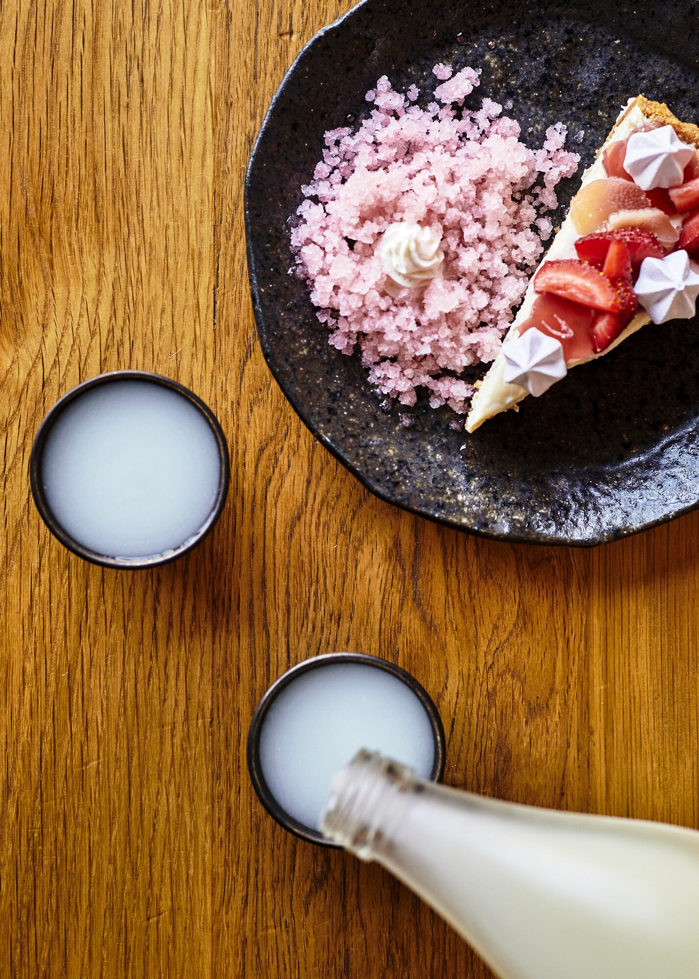 Cloudy nigori is poured in a cup beside a plate of ume cheesecake topped with strawberry rhubarb jellies, hibiscus meringue and red shiso granita.