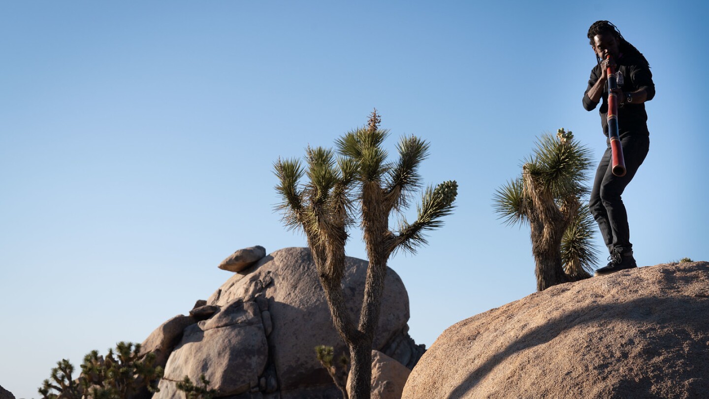 Kennedy Verrett stands on top of a rock formation, playing a didjeridu — a long, wind instrument. Behind him are other desert rock formations and Joshua trees set against a clear, blue desert sky.