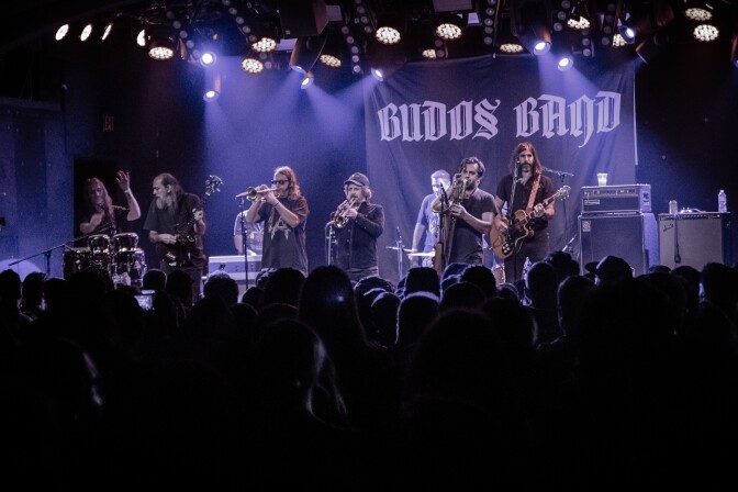 A band performs on stage lit in purple lighting. The band is composed of (from left to right) a percussionist, a bassist, two trumpet players, a saxophonist and a guitarist. Behind is a black banner that reads, "Budos Band" in stylized white typography. At the foreground of the photo is a silhouette of a densely packed crowd, outlined by the purple light coming from the stage.