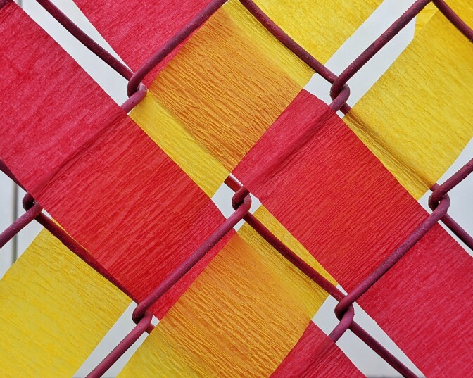 Pieces of red and yellow paper are woven in a pattern on a fence.