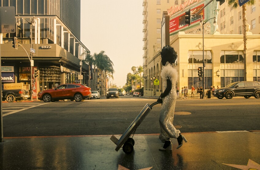 A person wearing a sparkly white dress over a black morph suit pushes a slab of concrete on a dolly down the Hollywood Walk of Fame. The person is wearing a glamorous early Hollywood wig and sparkly high heels.