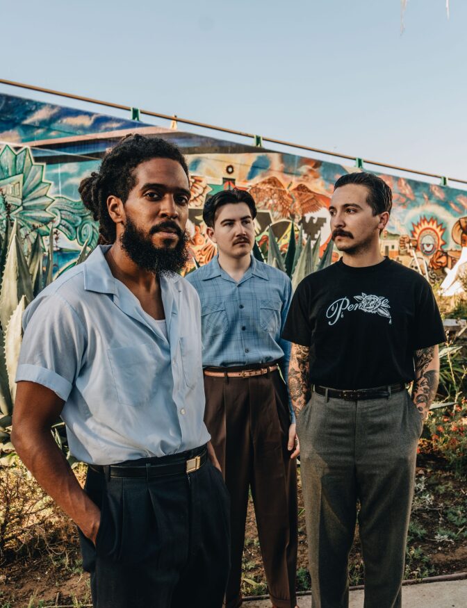 The members of Thee Sacred Soul, Sal Samano, Alex Garcia and Josh Lane, stand together in front of a mural.