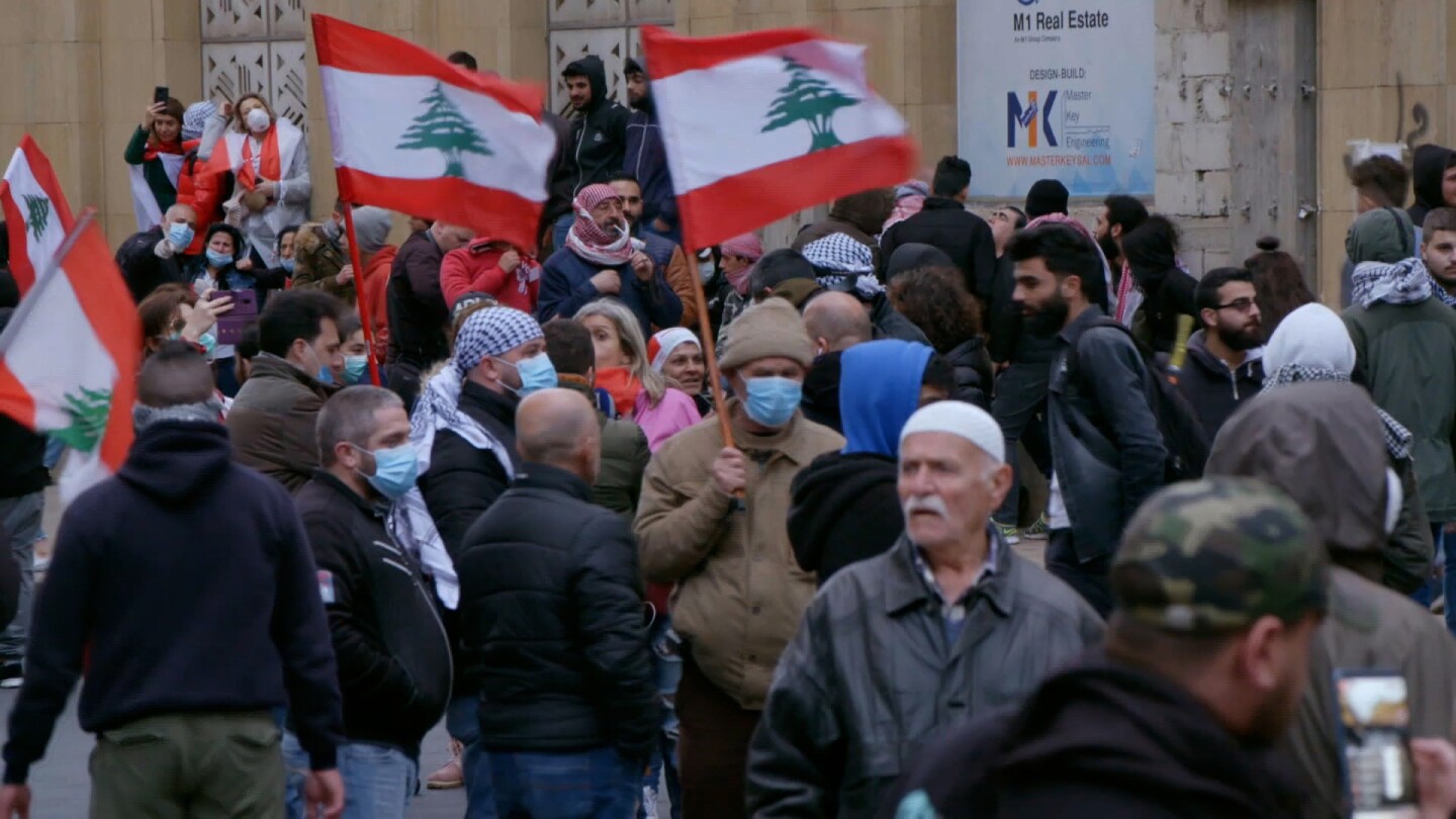 A crowd gathers on the street, some people holding up flags of Lebanon.