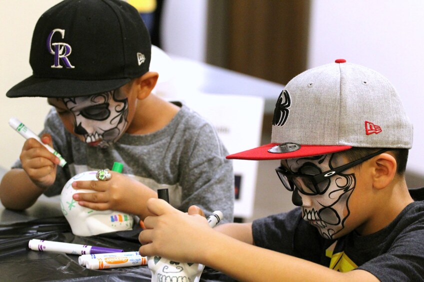 Two young boys wear skull face paint as they decorate small white plastic skulls for Day of the Dead.