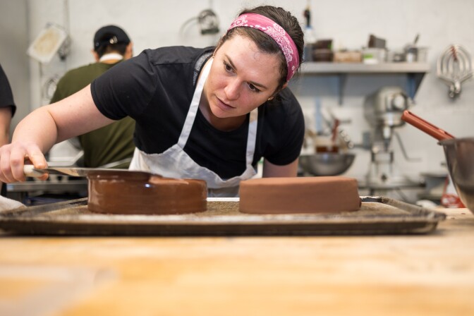 A woman wearing a white apron and a pink bandana around her head uses an offset spatula to glaze a chocolate cake.