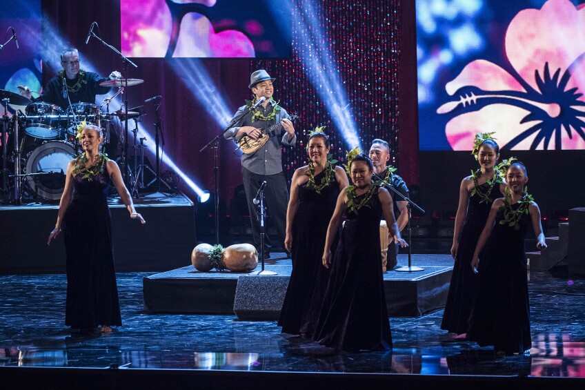 Daniel Ho and Halau Hula Keali'i o Nalani perform at 58th Annual LA County Holiday Celebration in Dorothy Chandler Pavilion. Daniel Ho is standing on a small square platform on stage as he plays the ukulele and sings into a microphone. In front of him on stage are five women wearing long black gowns, dancing. In the background, to the left is a man playing drums on a slightly elevated platform. Everyone on stage is wearing a lei made of green leaves around their neck, with the women also wearing green leaves in their hair which is slicked back into buns.