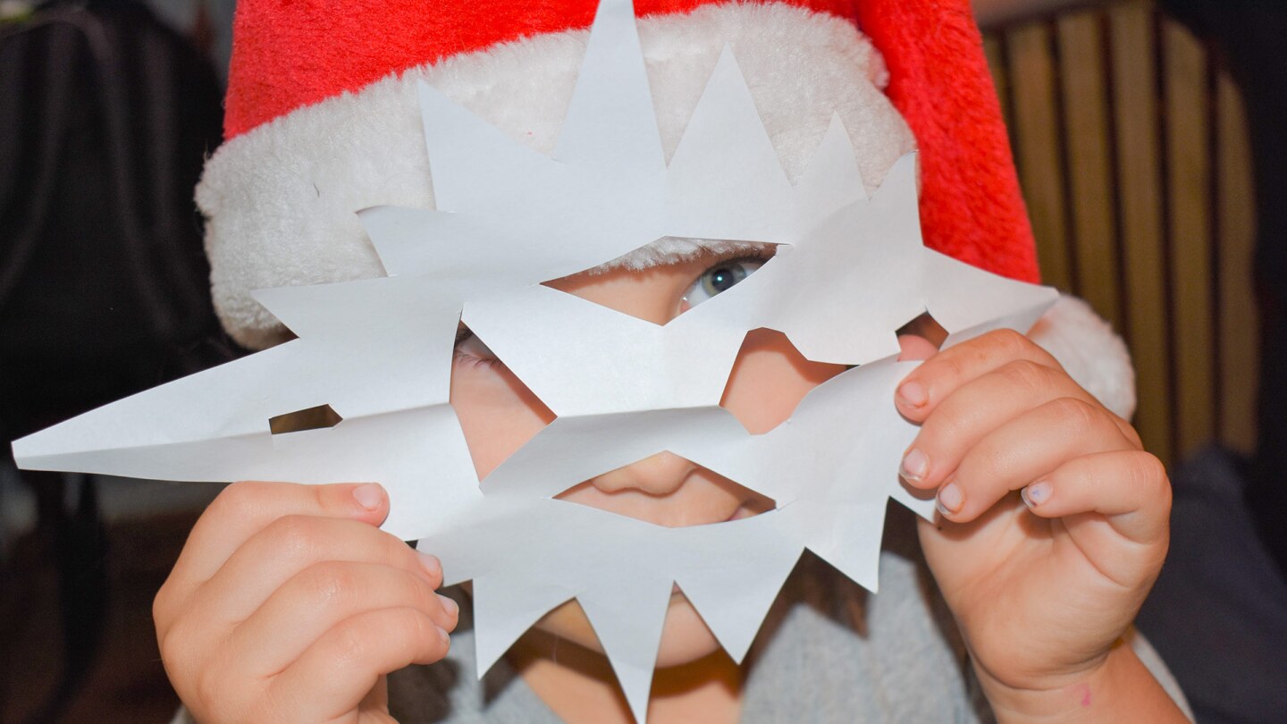 A small child wearing a red Santa hat holds a paper snowflake in front of the face like a mask.