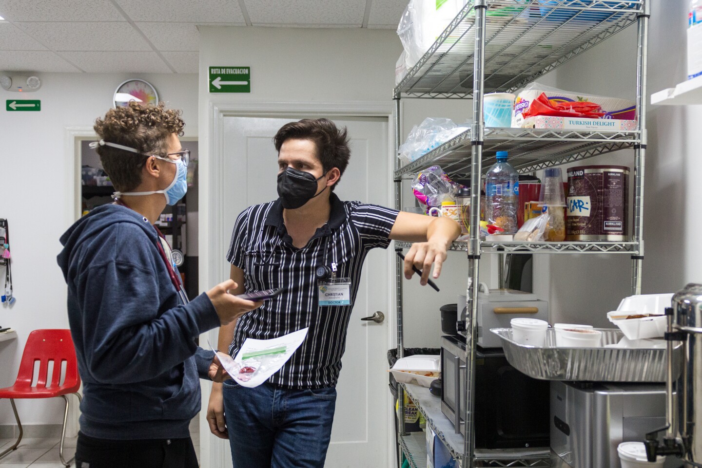 Two volunteers at the clinic talk to one another. One of the volunteers leans on a metal shelf with a microwave and various food supplies as they speak.