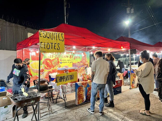 Customers are lined up in front of a red tent with a yellow hand-written sign that reads "Esquites" and "Elotes." On the table are two big, orange water jugs with a yellow sign in front of it reading, "Champurrado." Two people are working behind the table, grilling corn.