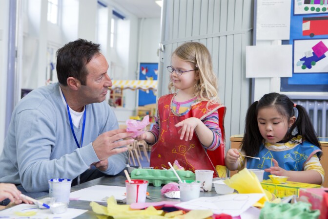Male teacher talking to one of his nursery students in the classroom. They are doing arts and crafts.