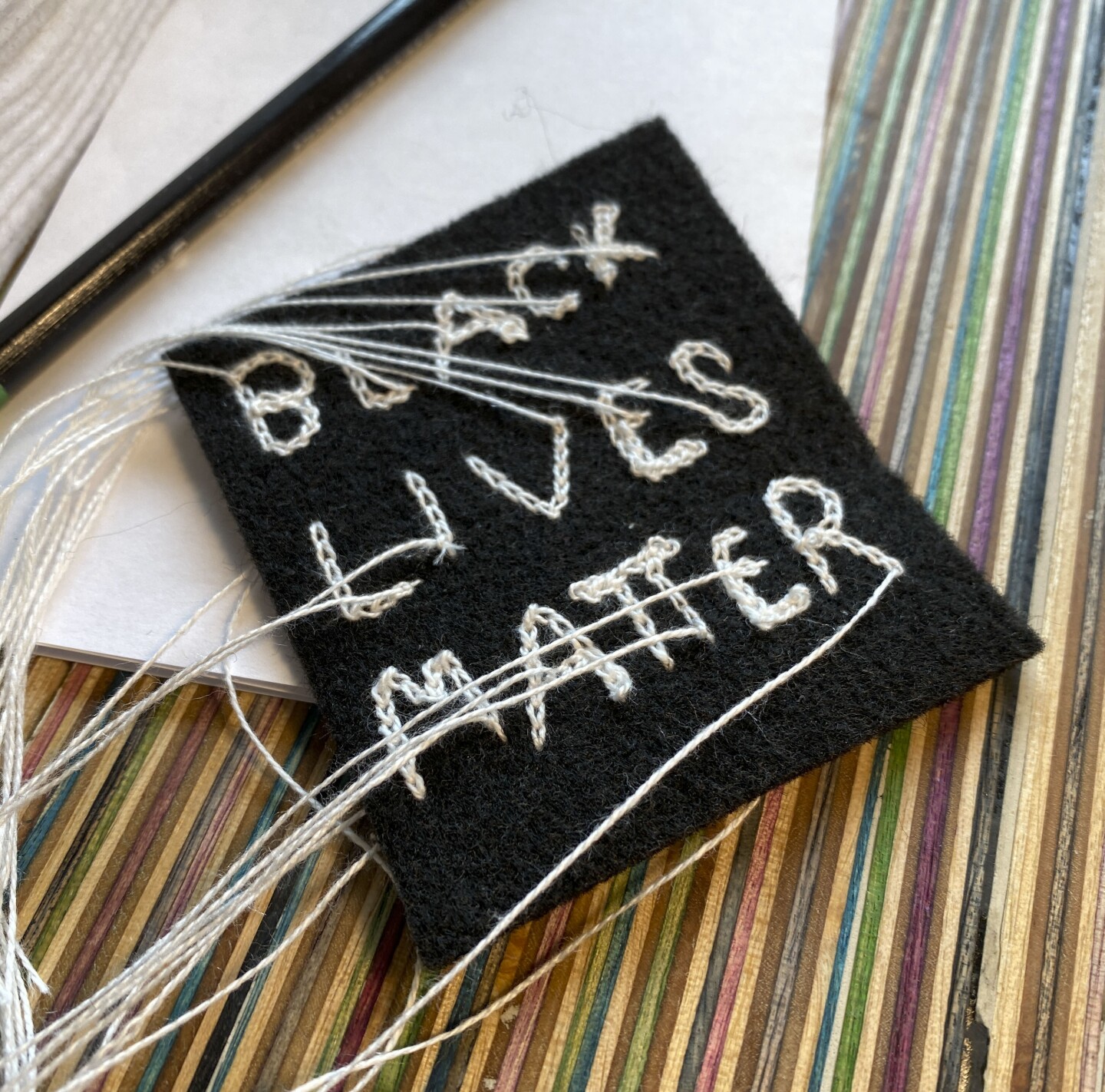 A black, square-shaped piece of fabric with chain stitch embroidery reading the words, "BLACK LIVES MATTER" in white thread. The ends of the threads are long and loose. The patch sits on top of a multicolored, striped surface.
