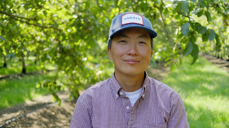 A profile photo of Kristyn Leach looking direct to camera. They're wearing a checkered button-up collared shirt and a blue cap that reads, "Sakata." Behind them are lines of green trees.