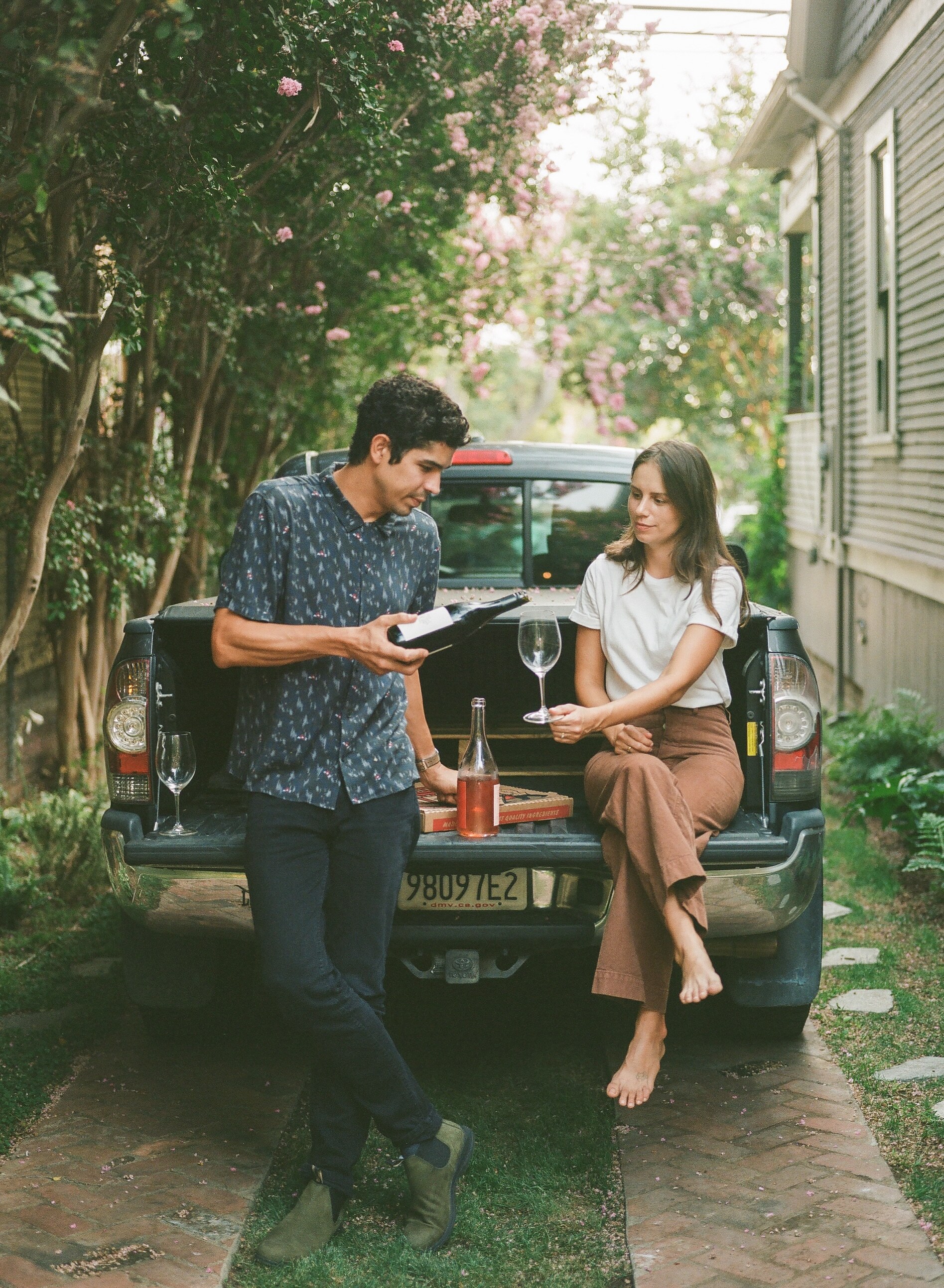 Rachel DeAscentiis sit on the bed of a pickup truck while Michael DeAscentiis leans against it. He's holding a bottle of wine, ready to pour it into a wine glass held out by Rachel.