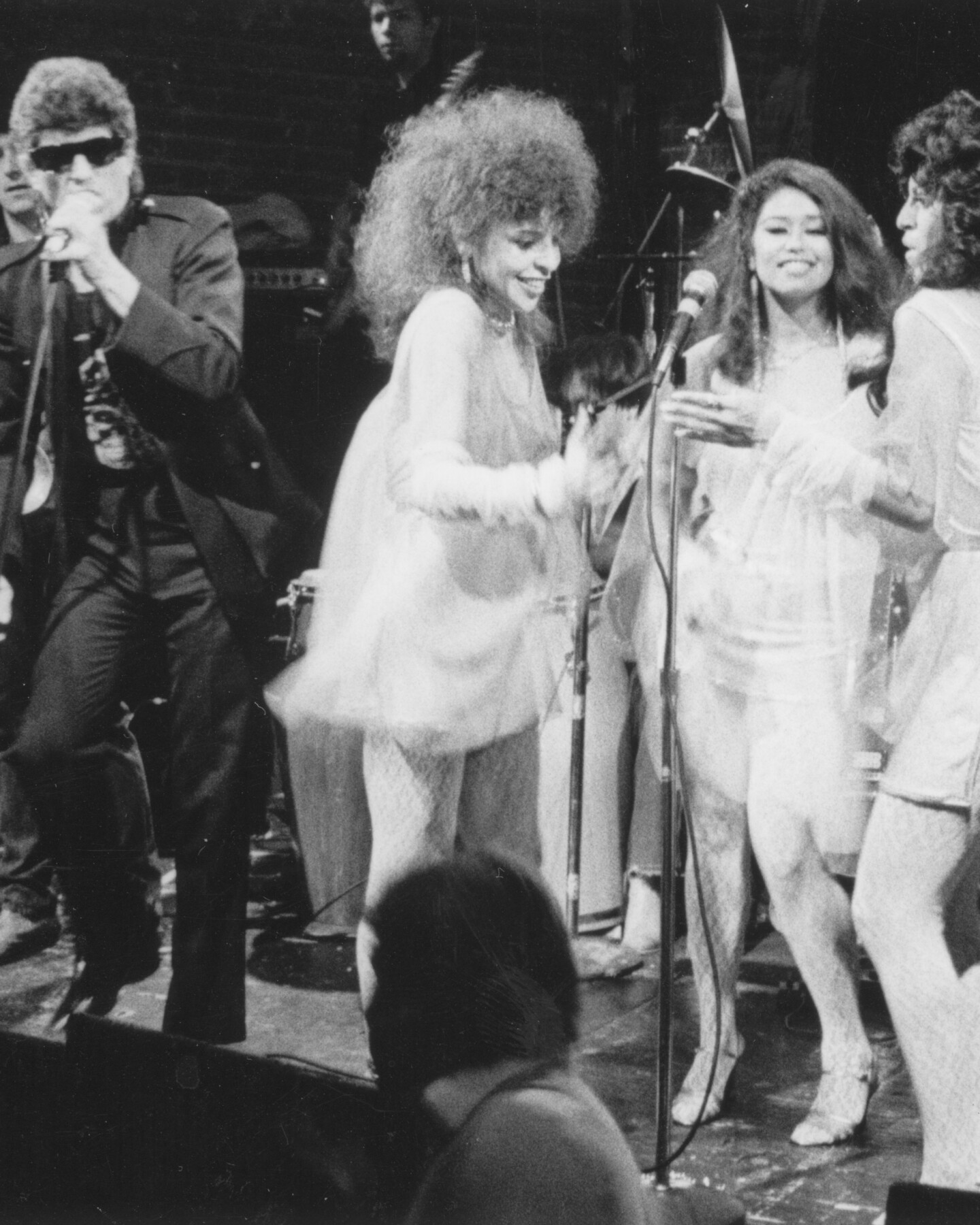 A man in sunglasses sings on stage with three women backing him up.