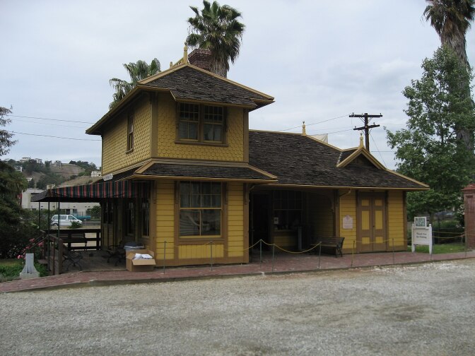 The Palms Depot, a mustard yellow Queen Anne with gingerbread cornices and brown trim, stands before a dirt path with trees sparsely surrounding it.