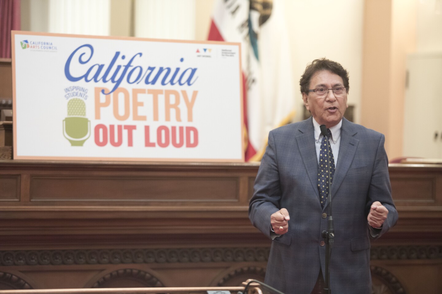 Larry Baza stands on the right-hand side of the photo in a dark gray suit and black and white polka dotted tie. He is speaking into a microphone indoors. Behind him is a banner that reads, "California Poetry Out Loud."