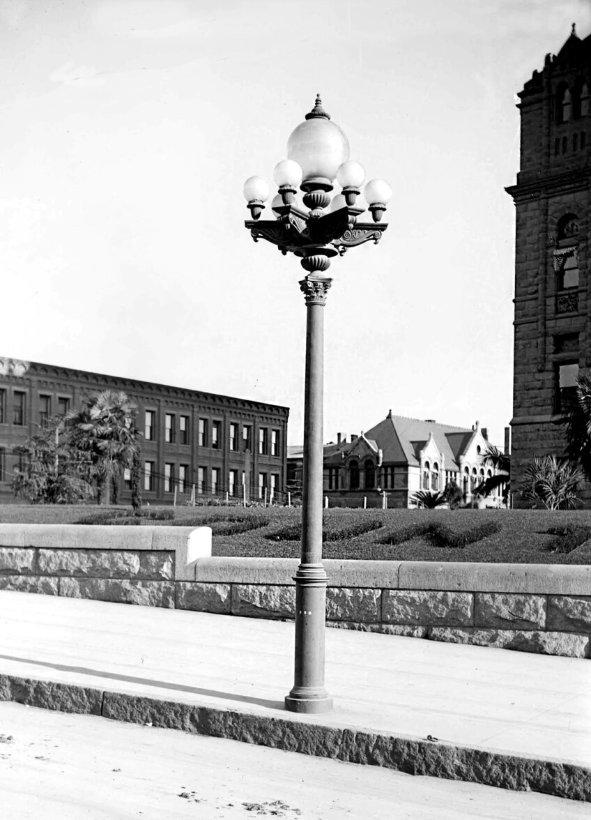 A black and white archival photo of a lamp post on the side of the road. The lamp post features six circular bulbs surrounding a larger globe bulb.