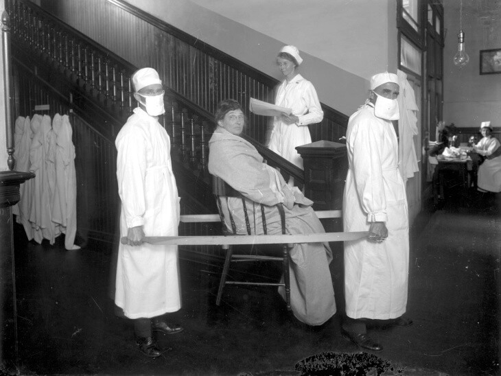 Pasadena, approximately 1919: Two orderlies wearing breathing masks carry a patient in a chair, possibly during the influenza epidemic of 1918 and 1919. | Harold A. Parker/Huntington Digital Library