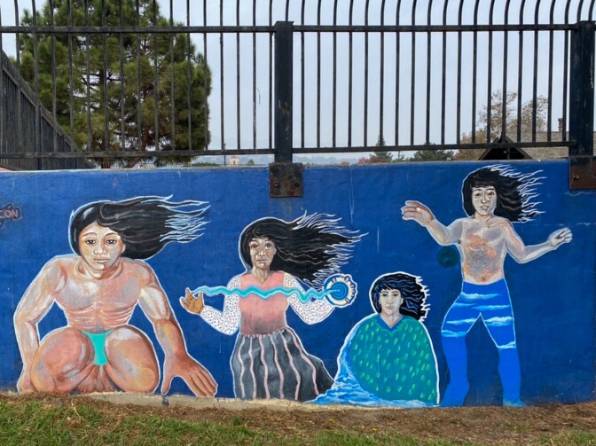 A mural is painted on a concrete half-wall. Above, black steel bar fencing is bolted onto the wall. The mural depicts Chumash imagery, featuring four Indigenous people with long Black hair flowing in the wind.