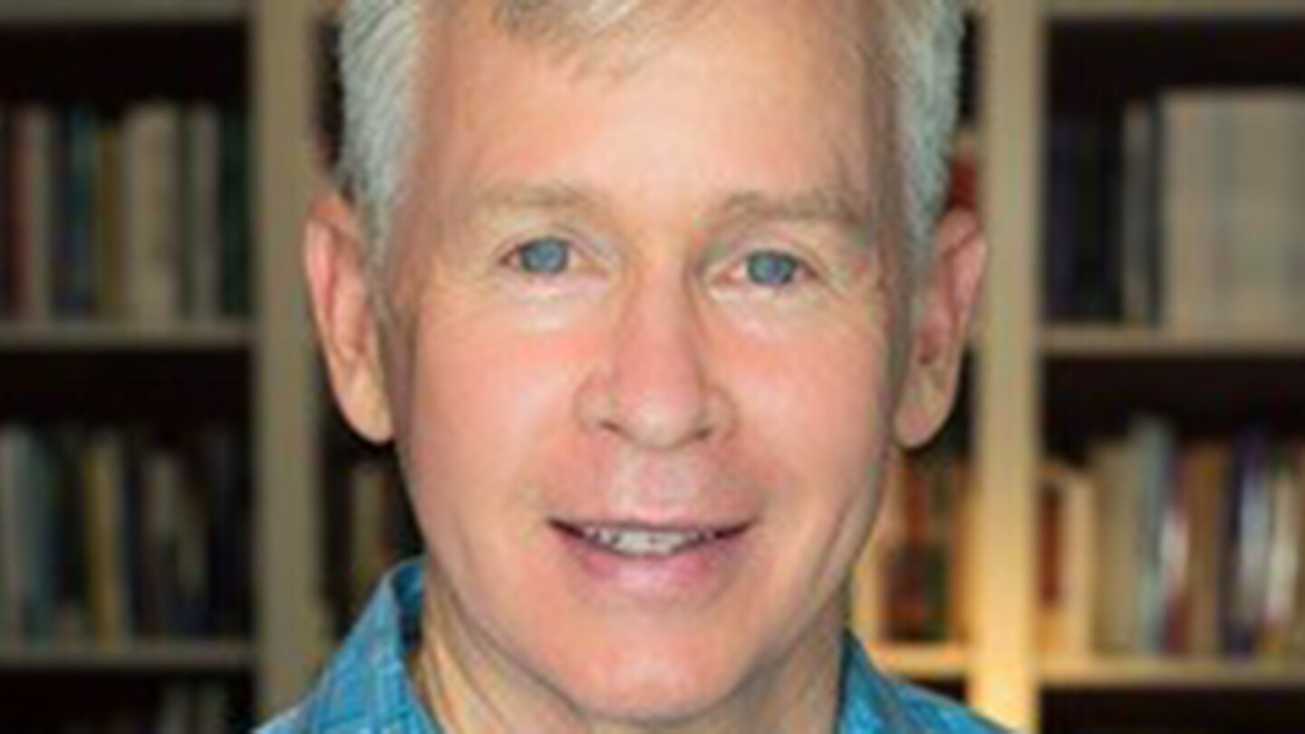 Dr. Philip Lance wears his white hair short. He is standing in front of a bookshelf and is wearing a blue plaid button down.