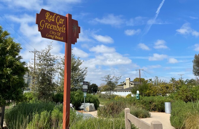 A photo of a red wooden sign that reads, "Red Car Greenbelt" in yellow paint. Below that reads, "City of Long Beach" in smaller, yellow lettering. The sign stands against a bright blue sky and amidst a lush green space.