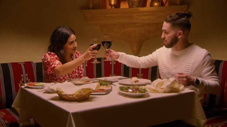 Natasha, who is wearing a red dress with white flowers, and Shant, who is wearing a white, cable-knit sweater, clink wine glasses over a dinner table topped with a white tablecloth and various dishes. Natasha and Shant look at each other as they toast.