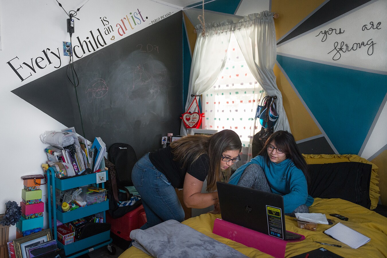 Rocio helps her daughter, Rhianna, with online classes. Rhianna sits on her bed, topped with a mustard yellow comforter and her laptop opened in front of her. The walls of her room are painted in a vibrant, geometric pattern. Along her wall are various school supplies, books and other miscellaneous items.