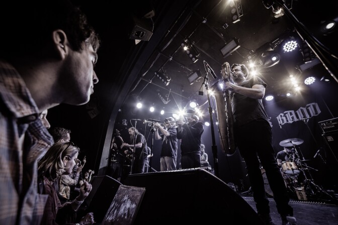 A band performs on a stage. From left to right there is a guitarist, two trumpet players and a saxophonist. The audience listens to the left of the image. Behind the band are bright white stage lights.