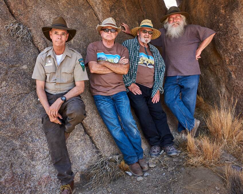 A group photo of four older men standing in crevice in a rock formation. The man on the far left is wearing a park ranger outfit. Next to him a man wearing a wide-brimmed hat and a brown t-shirt stands with his arms crossed. Next to him, a man wears a straw, wide-brim hat and a green plaid button up over a brown t-shirt. The man on the far right is wearing a wide-brimmed hat and a brown t-shirt. He stands with his hand on his hip.