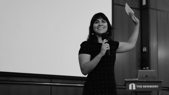 Black and white photo of a woman speaking into a microphone with her left hand raised holding a piece of paper