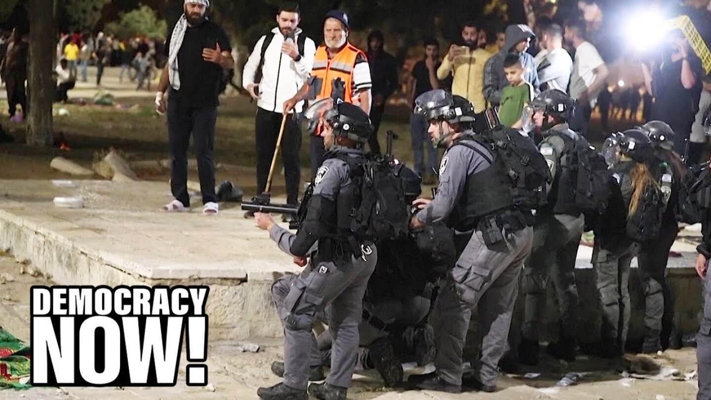 Armed policemen have their weapons at the ready while a crowd of people watch.