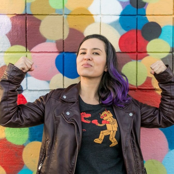 A woman in a black and purple hair and a black leather jacket flexes her biceps in front of a colorful wall.