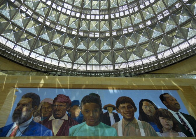 A mural painting features various people of different genders, ages, ethnicities and races. Above the mural is an elaborate art deco-style glass dome, reflecting geometric lights shapes onto the mural.